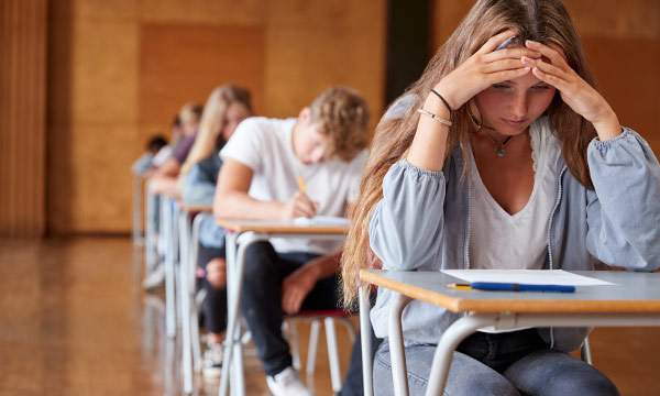 Gestresster Schüler am Schreibtisch während einer Prüfung mit anderen Schülern im Hintergrund